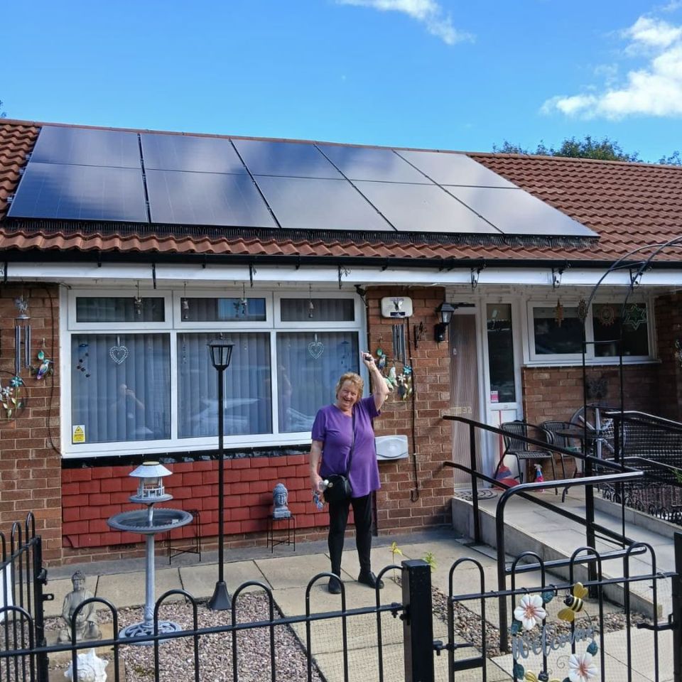 A woman stands in front of a house with solar panels on the roof, smiling and raising a fist in the air. A woman stands in front of a house with solar panels on the roof, smiling and raising a fist in the air.